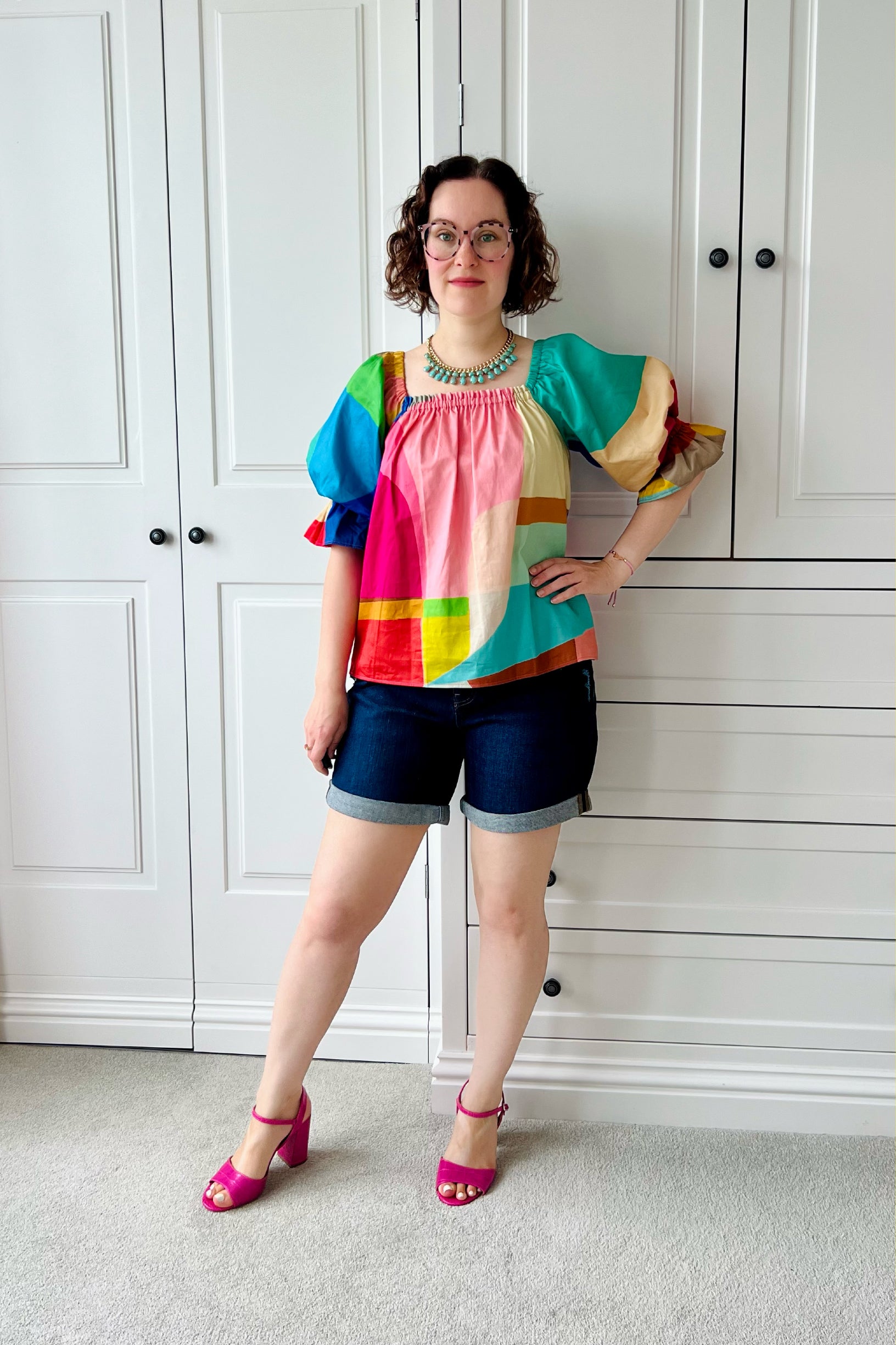 Woman wearing a colorful blouse and shorts standing in front of a white wardrobe. The sleeves are puffy and make a statement. Easy sewing pattern for beginners.