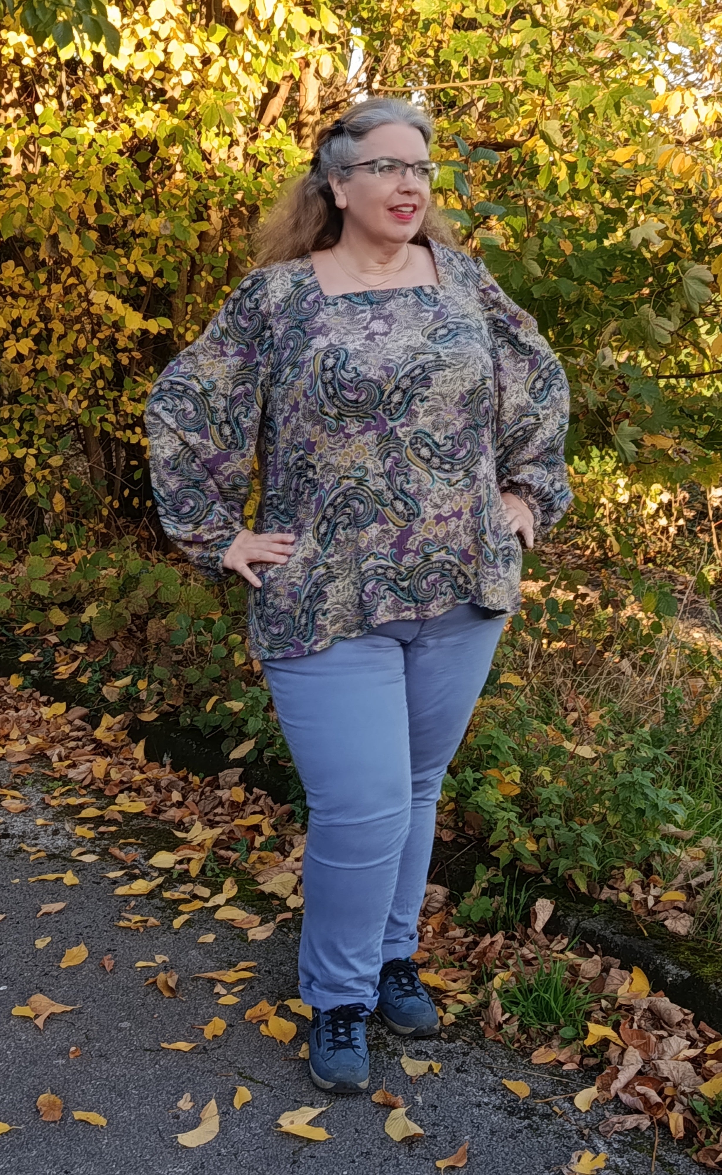 Woman wearing the Zara blouse in a patterned lightweight woven fabric and blue jeans standing outdoors with trees and fallen leaves in the background