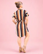 A woman wearing a short-sleeve belted dress with vertical stripes in orange, brown, black, and cream, photographed from the back against a solid pink studio background.