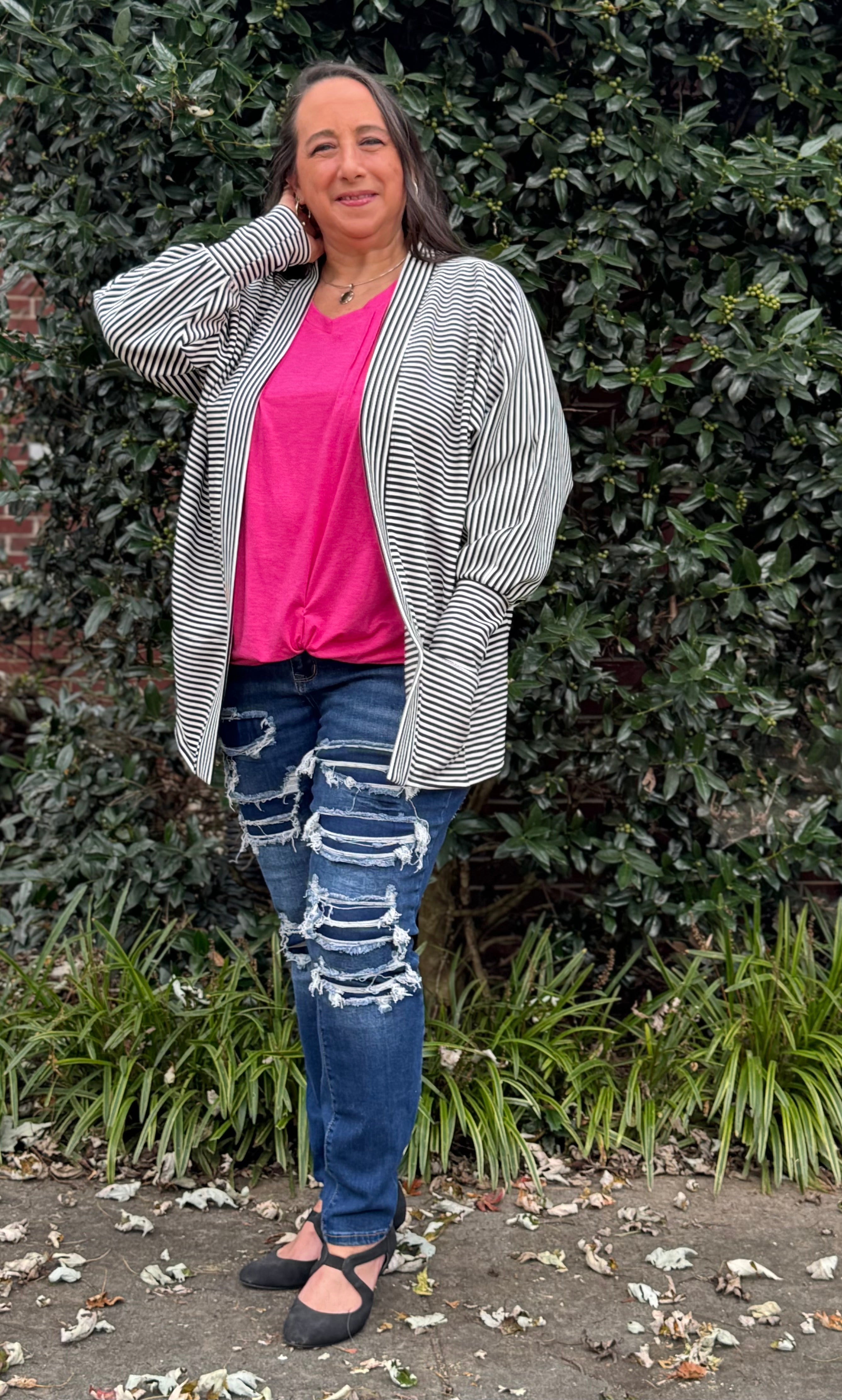 Woman wearing a striped cardigan, pink shirt, and ripped jeans standing against a green bush. PDF sewing pattern.