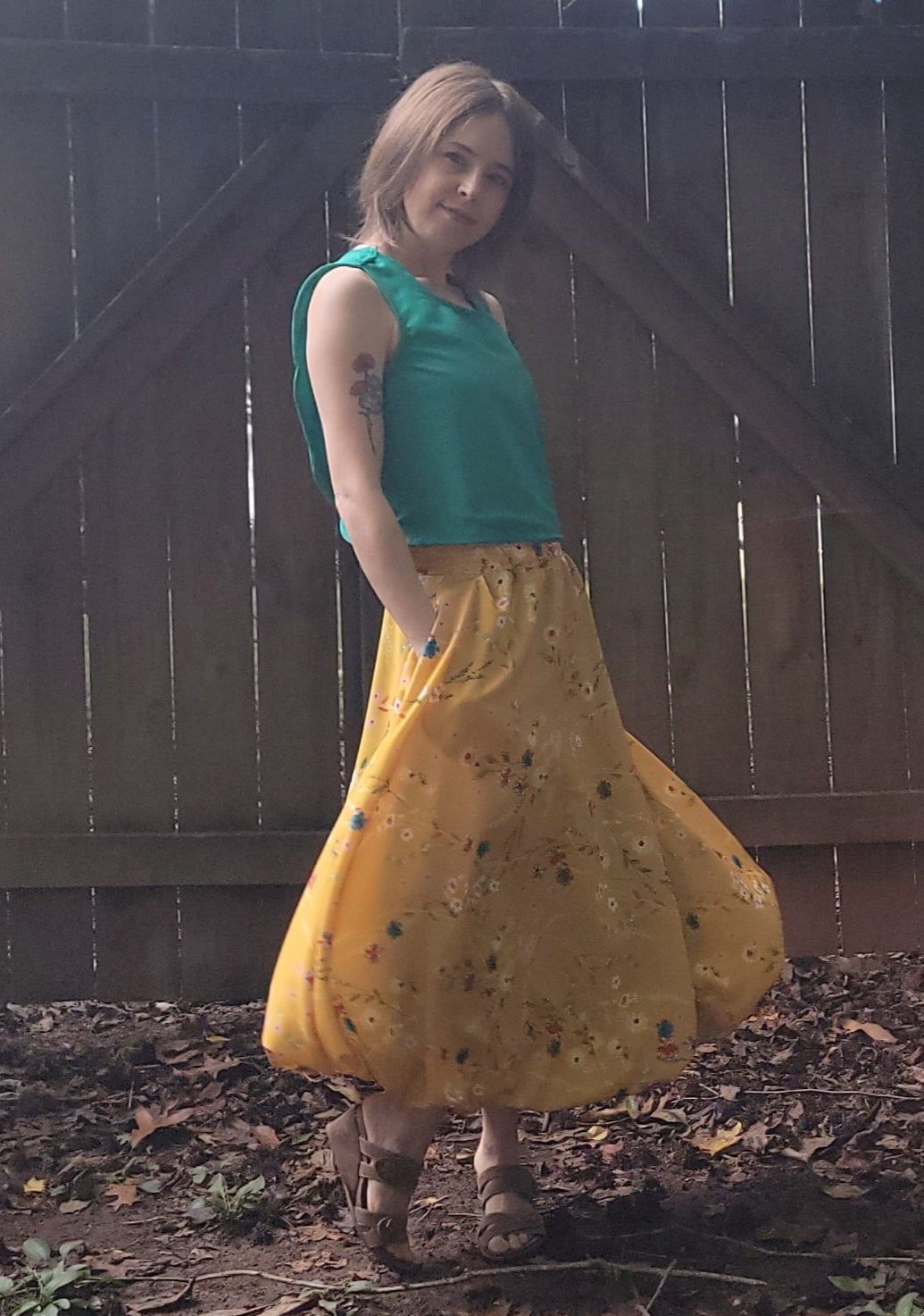 Woman wearing a green top and yellow skirt standing in front of a wooden fence with trees in the background.