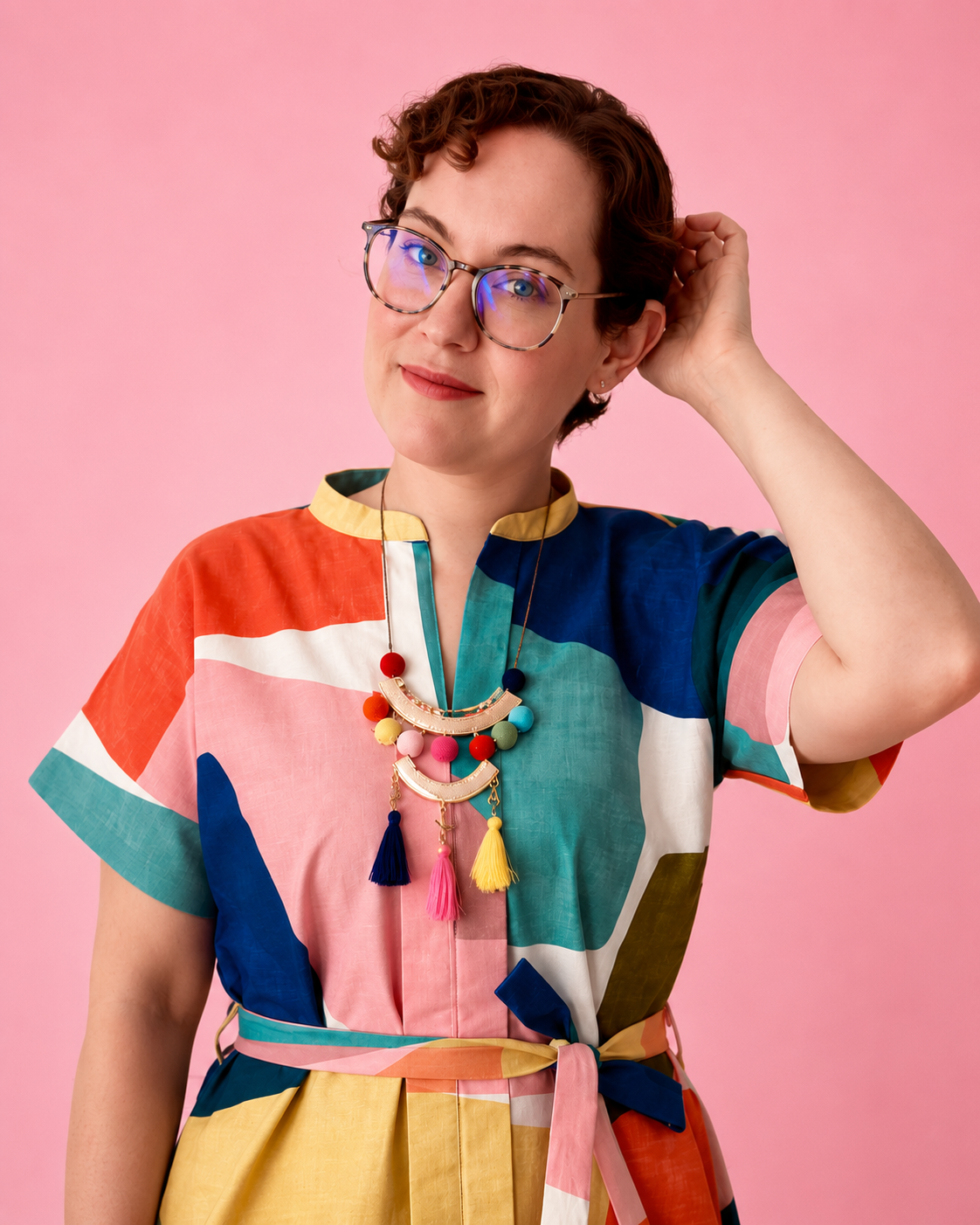 A woman with short curly brown hair and glasses wearing a colorful geometric print dress and matching sash, posed against a solid pink background. She made this beautiful dress using Ellie and Mac PDF sewing patterns. They include A0, A4, Letter, print files and projector sewing file. They also include a full sew along video tutorial.