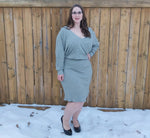 Woman wearing a grey crossover dolman sweater dress standing in front of a wooden fence with snow on the ground.
