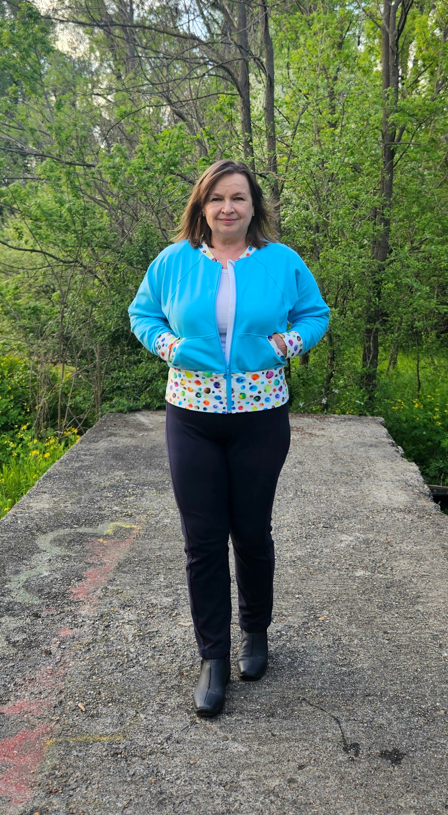 Woman on a path modeling a aqua and blue athletic bomber jacket with zip closure, kangaroo pocket detail, and cuffs. Easy pdf sewing pattern.