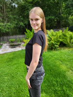 Teen girl standing on grass with trees in the background modeling from the side, a black dolman t-shirt.