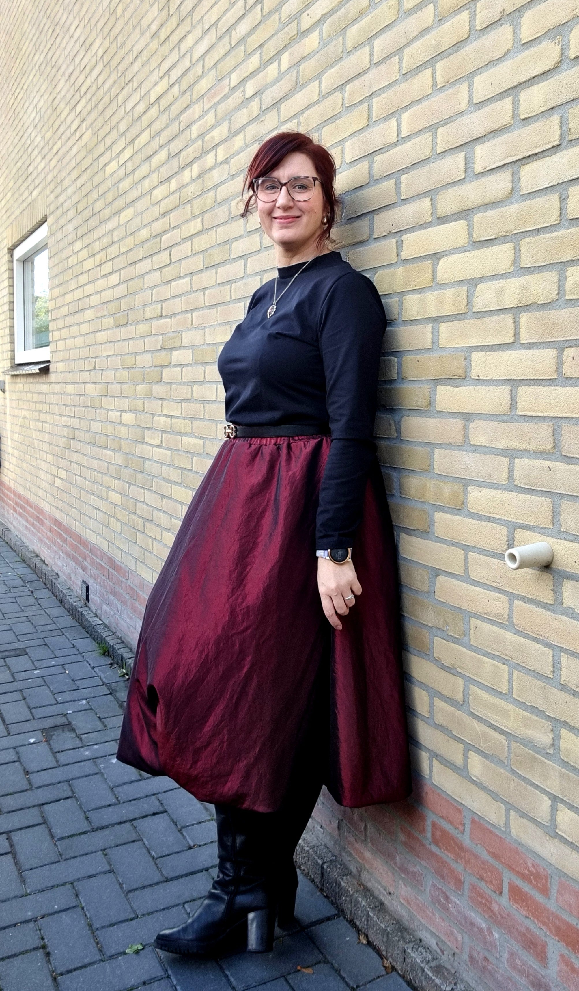 Person wearing a black top and burgundy skirt leaning against a brick wall.