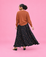 A woman is shown from behind wearing a rust-colored knit sweater and a long black skirt with a small floral print, standing against a solid pink studio background.