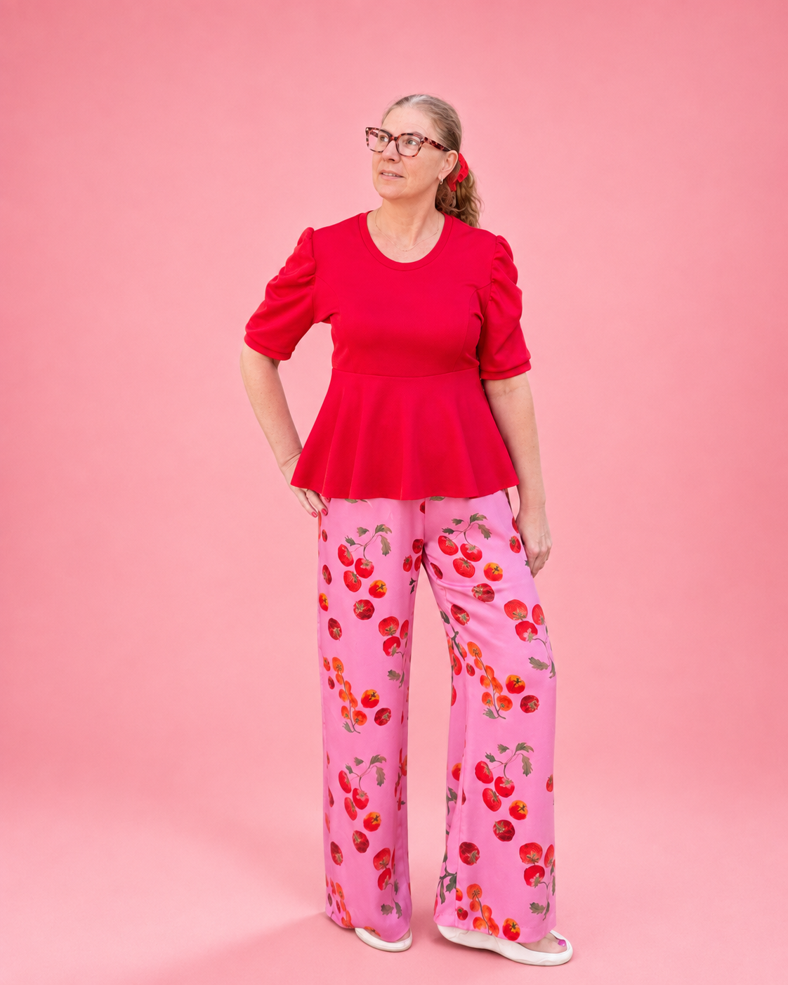 A woman modeling a bright red short-sleeve peplum top and pink wide-leg pants with a red and orange cherry print, standing against a solid pink background. The top is made using Ellie and Mac PDF sewing patterns. They include A4, Letter, A0, and Projector pattern files. They also include a full sew-along tutorial. 