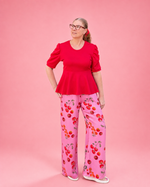 A woman modeling a bright red short-sleeve peplum top and pink wide-leg pants with a red and orange cherry print, standing against a solid pink background. The top is made using Ellie and Mac PDF sewing patterns. They include A4, Letter, A0, and Projector pattern files. They also include a full sew-along tutorial. 
