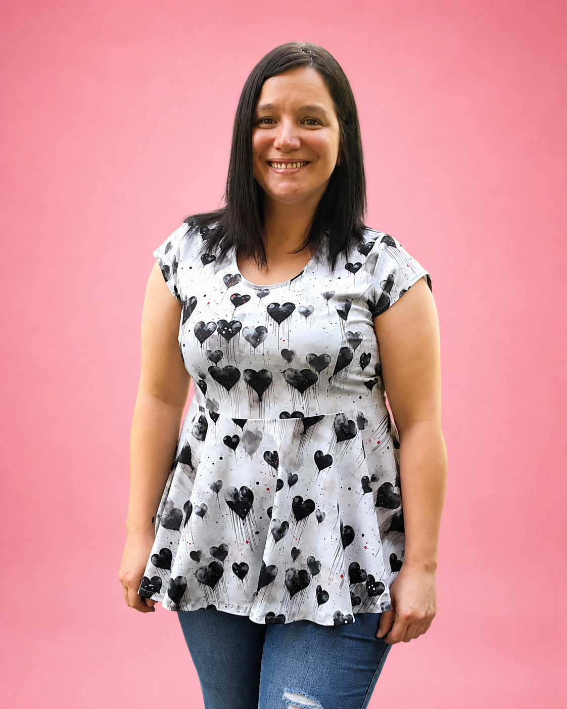 A woman with dark hair smiling, wearing a white short-sleeve peplum top with a black heart print, paired with blue jeans, set against a solid pink background. The top is made using Ellie and Mac PDF sewing patterns. They include A4, Letter, A0, and Projector pattern files. They also include a full sew-along tutorial. 
