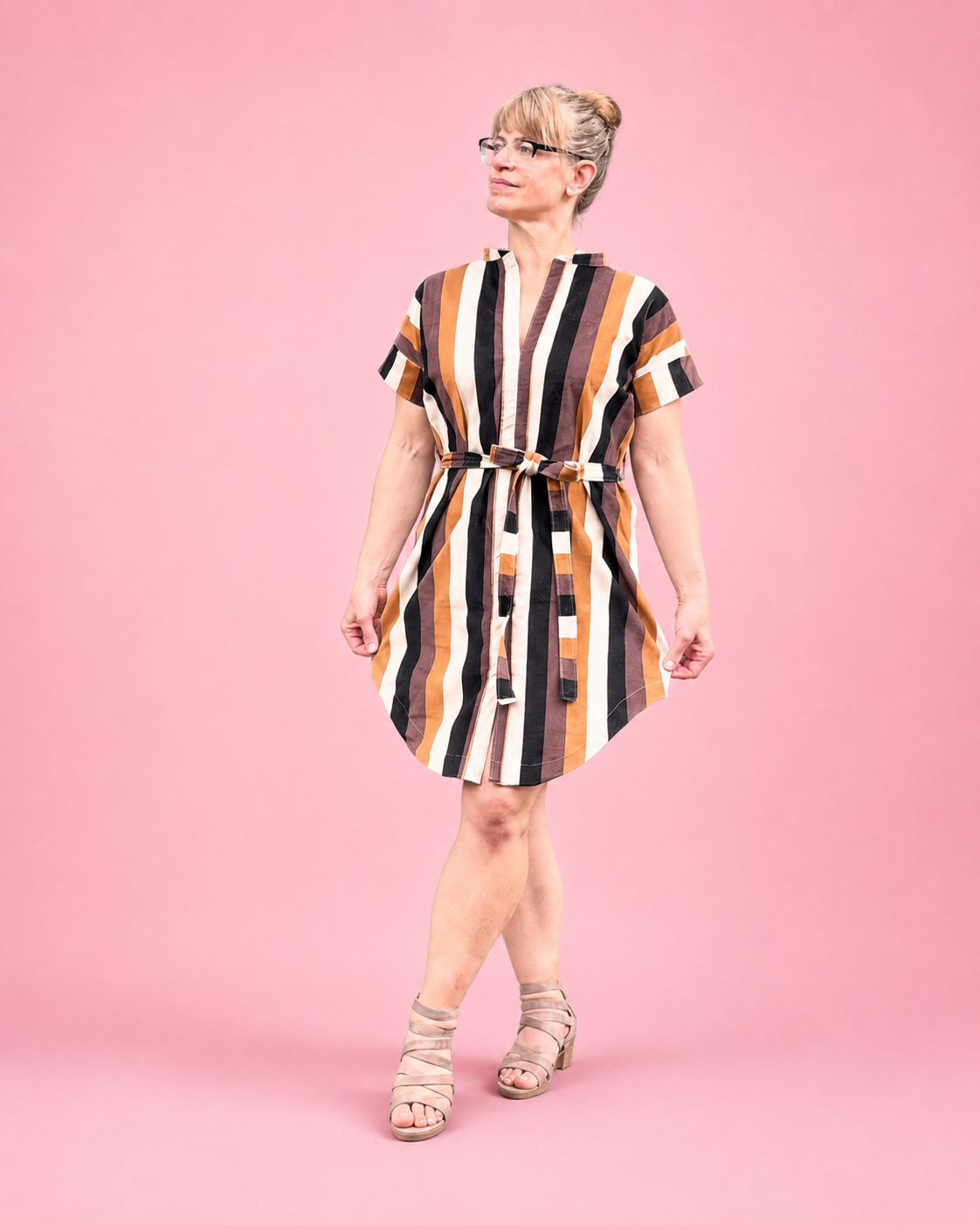 A woman modeling a short-sleeve mini dress with vertical brown, black, white, and tan stripes, featuring a matching fabric belt at the waist, posed against a solid pink background. Easy woven dress sewing pattern for women.