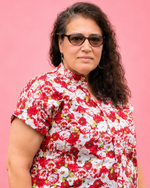 A woman with curly dark hair and sunglasses wearing a red and white floral print short-sleeve shirt, standing against a solid pink background. Blouse made using Ellie and Mac PDF sewing patterns. They include A0, A4, Letter, print files and projector sewing file. They also include a full sew along video tutorial.