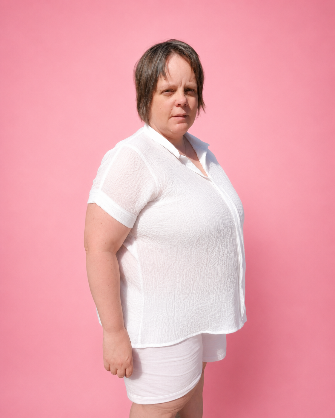 A woman modeling a white crinkle short-sleeve collared shirt and matching white shorts against a solid pink studio background.