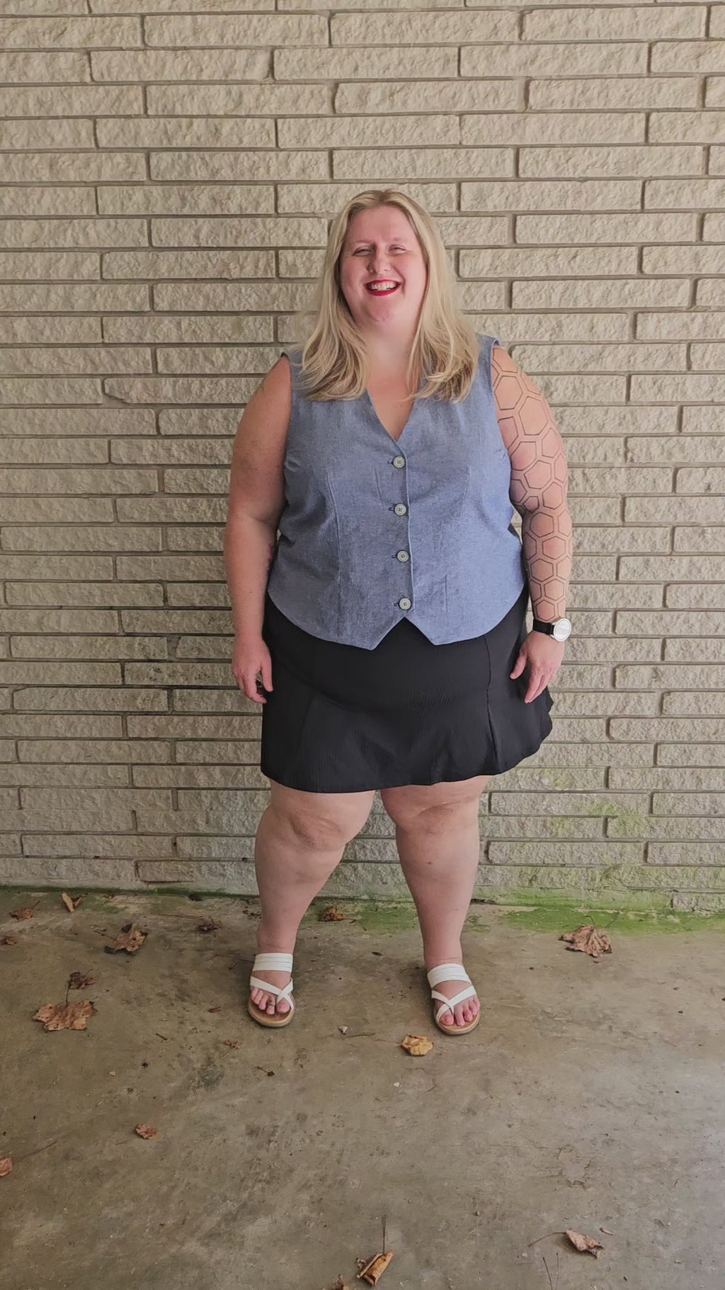 Woman wearing a denim vest and black mini skirt standing by a brick wall outside. The vest is the high hip length option made from the Veronik Waistcoat Vest sewing pattern by Ellie and Mac Sewing Patterns. 