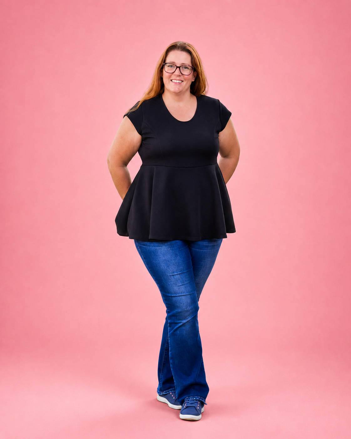 A woman modeling a black short-sleeve peplum top with blue jeans and blue sneakers, standing against a solid pink background. The top is made using Ellie and Mac PDF sewing patterns. They include A4, Letter, A0, and Projector pattern files. They also include a full sew-along tutorial. 