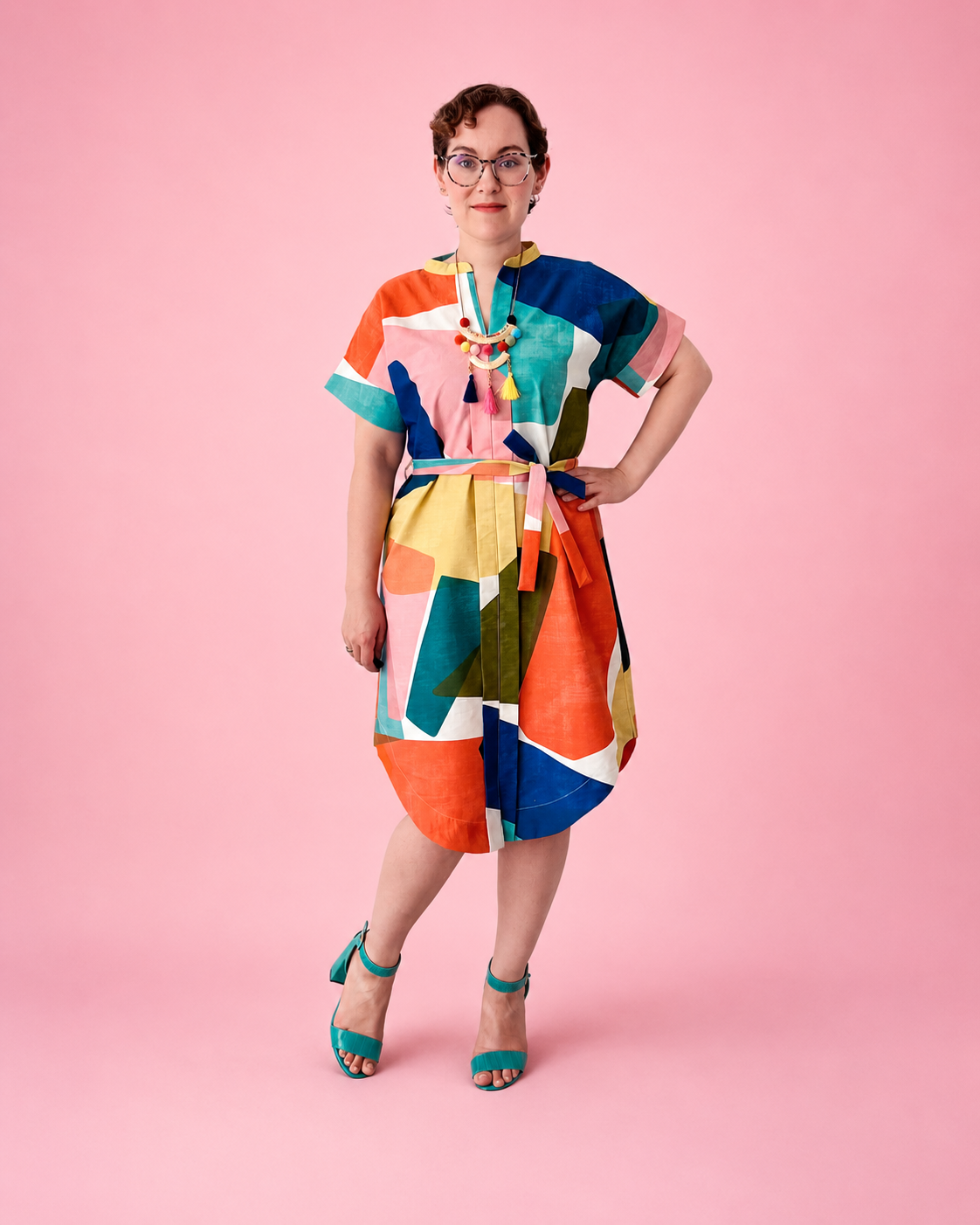 A woman wearing a colorful geometric print dress and teal heeled sandals, standing against a solid pink background. Dolman woven dress made using Ellie and Mac PDF sewing patterns. They include A0, A4, Letter, print files and projector sewing file. They also include a full sew along video tutorial.
