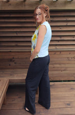 Woman wearing a floral top and black pants standing against a wooden wall. 