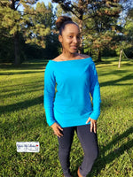 Women hair in a high ponytail wearing a teal off shoulder sweater with banded neckline standing in a park.
