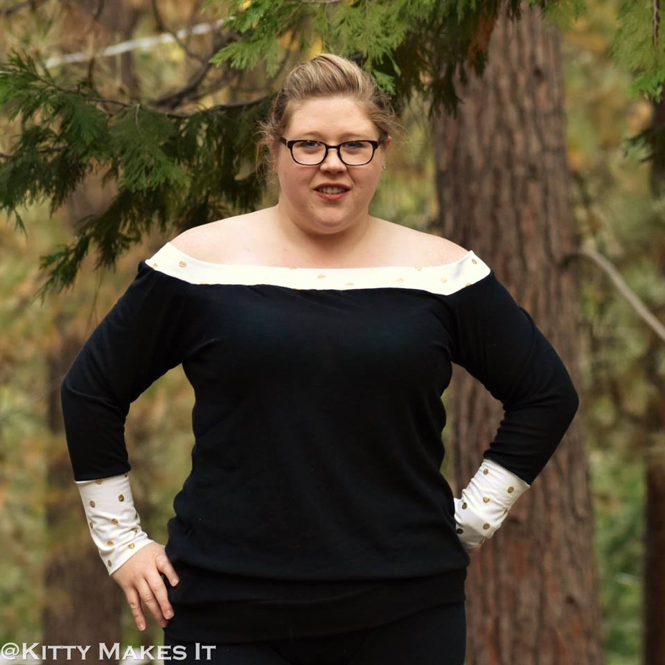 Woman wearing a black sweater with banded off shoulder neckline and tall cuffs.