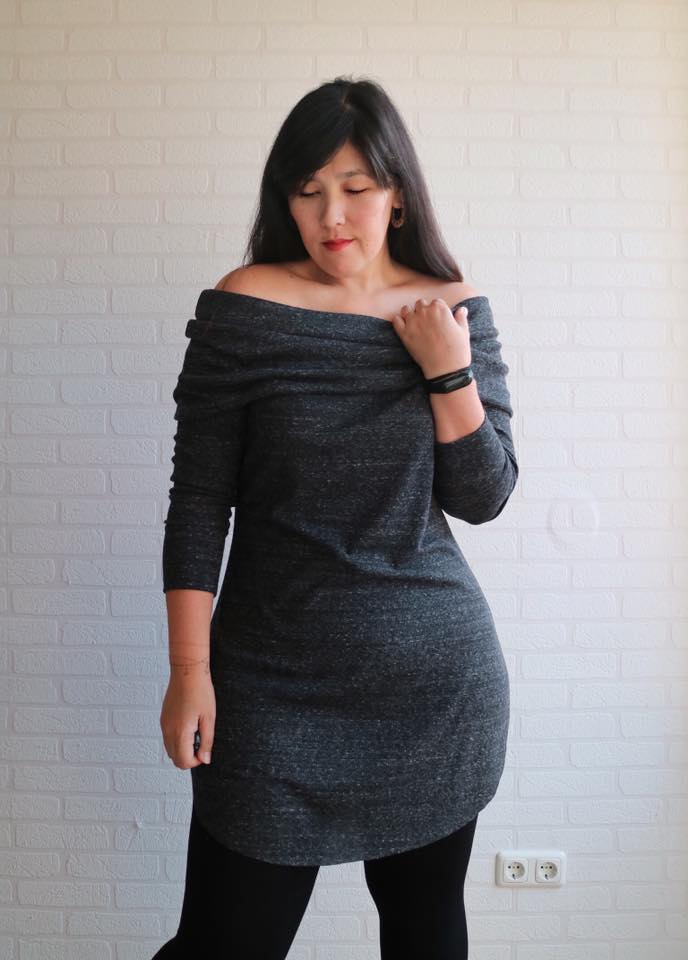 A woman with dark hair standing in front of a white wall wearing a grey sweater dress with slouchy off shoulder cowl neck. Dress has a curved hemline and she is wearing black tights.