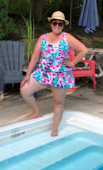 Ellie & Mac Gathered Tankini Swimsuit Sewing Pattern. The image shows a woman modeling the finished swimsuit in a vibrant floral abstract print, alongside a pool. This PDF pattern is available in sizes XXS-6XL and printable on A4, A0, and Letter paper sizes. Projector file and video tutorial included. 