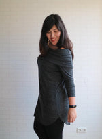 Woman standing in front of white brick wall with dark long hair, wearing a grey, soft knit sweater with curved hem detail and off shoulder slouchy cowl.