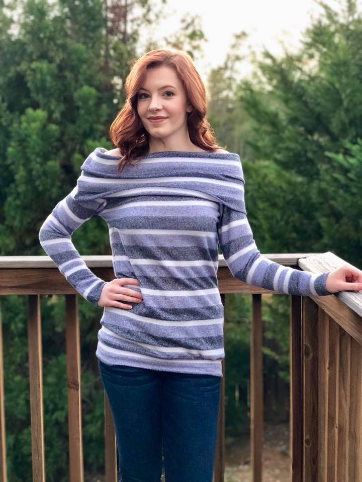 Women with red hair wearing a striped off shoulder sweater with slouchy cowl standing on a deck with trees in the background.