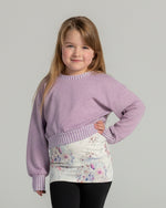 Young girl wearing a purple sweater and patterned skirt on a plain background