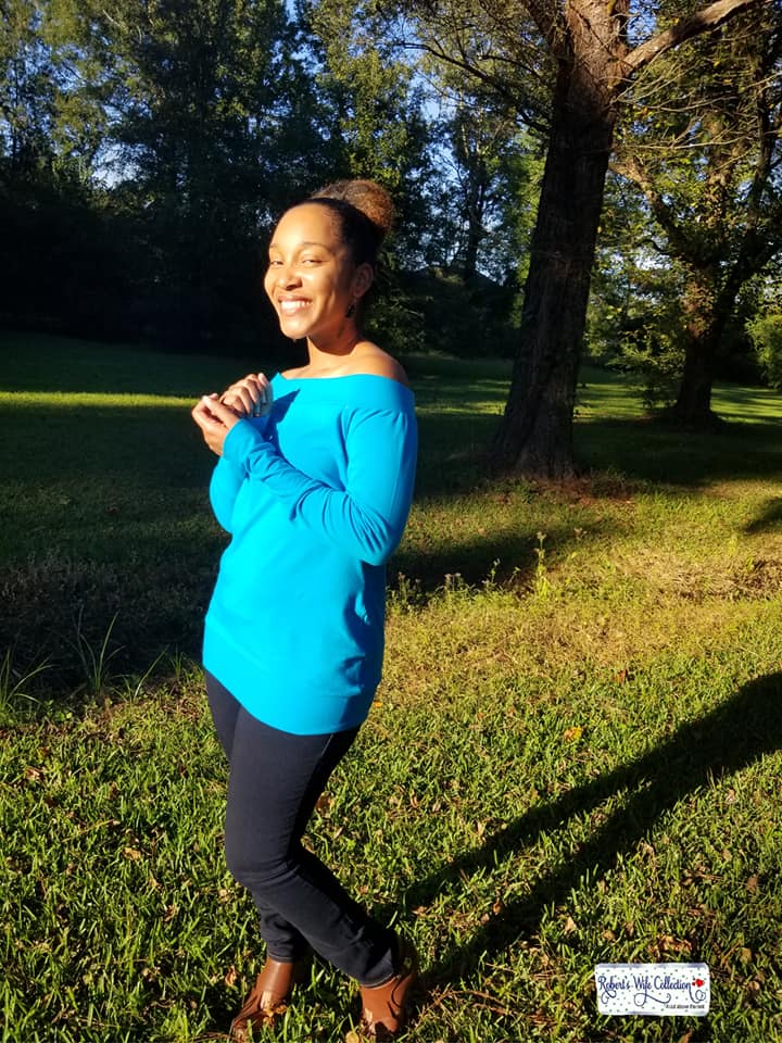 Women hair in a high ponytail wearing a teal off shoulder sweater with banded neckline standing in a park.