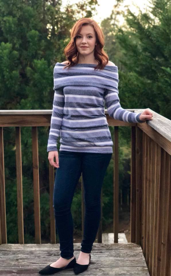 Women with red hair wearing a striped off shoulder sweater with slouchy cowl standing on a deck with trees in the background.