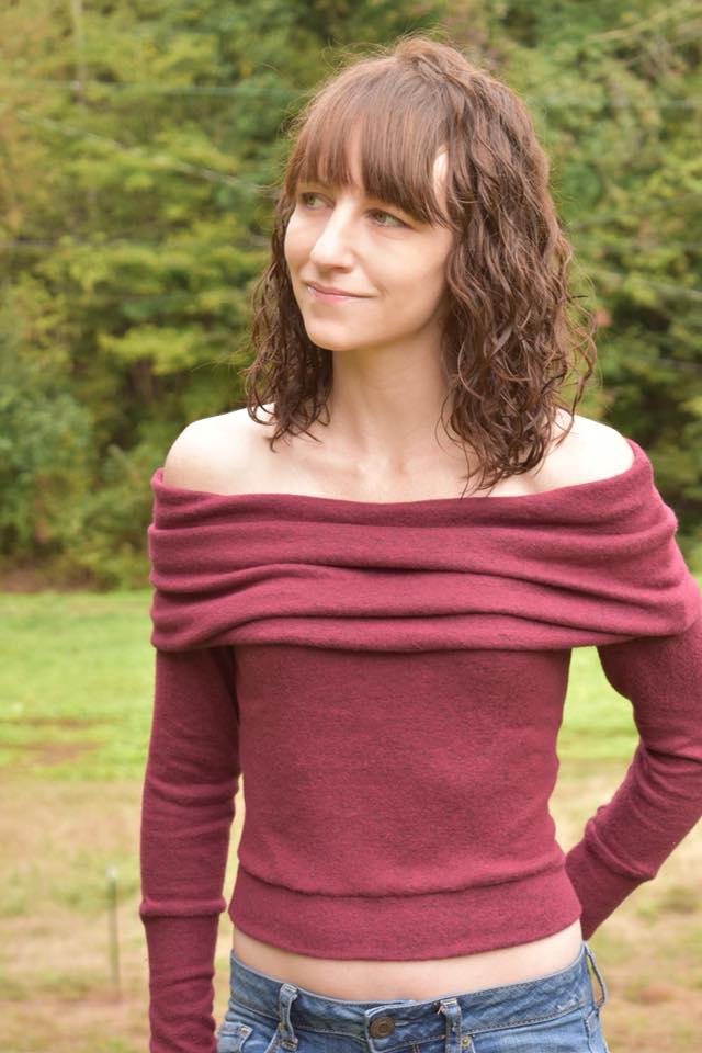 A woman with redish brown hair standing outside with trees in the background, wearing a red lightweight sweater with off-shoulder cowl and long cuffed sleeves. Hem of the crop sweater is banded.