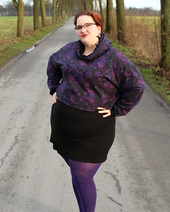 Woman wearing a purple sweater with cowl neck paired with a black skirt and purple stockings. 