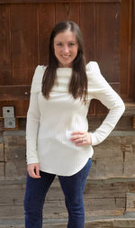 A woman with straight brown hair standing outside of a cabin, leaning on a railing, wearing a gorgeous white lightweight sweater with off-shoulder cowl and long cuffed sleeves. Hem of the sweater is hemmed and curved. Made using Ellie and Mac Sewing Patterns.
