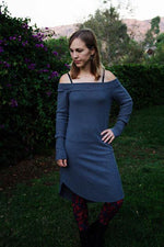 Woman outside by a garden, wearing a blue ribbed sweater knit dress that is knee length and long sleeve. Off shoulder banded neckline. PDF sewing pattern.