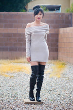Woman in a sexy tan mini sweater dress with long sleeves and oversized cowl off shoulder neckline. She is wearing black thigh high boots. 