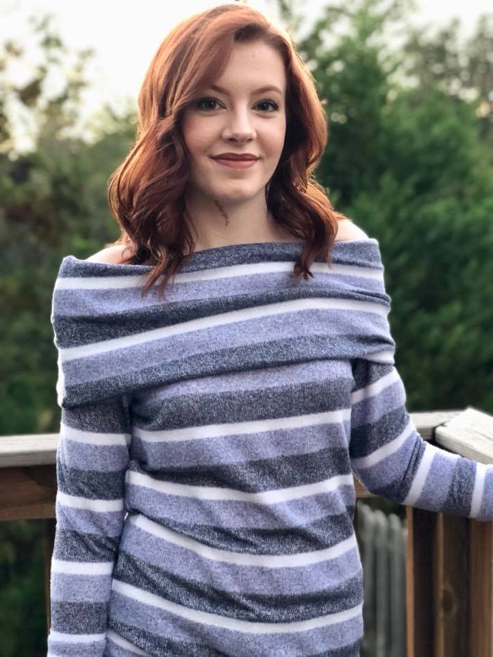 Women with red hair wearing a striped off shoulder sweater with slouchy cowl standing on a deck with trees in the background.