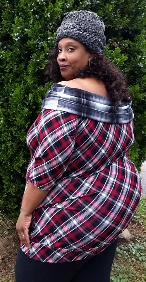 Woman in front of a tree wearing a beanie hat over her curly brown hair. The plaid sweater is tunic length and she is wearing black leggings underneath. The off shoulder cowl is in a contrasting fabric.