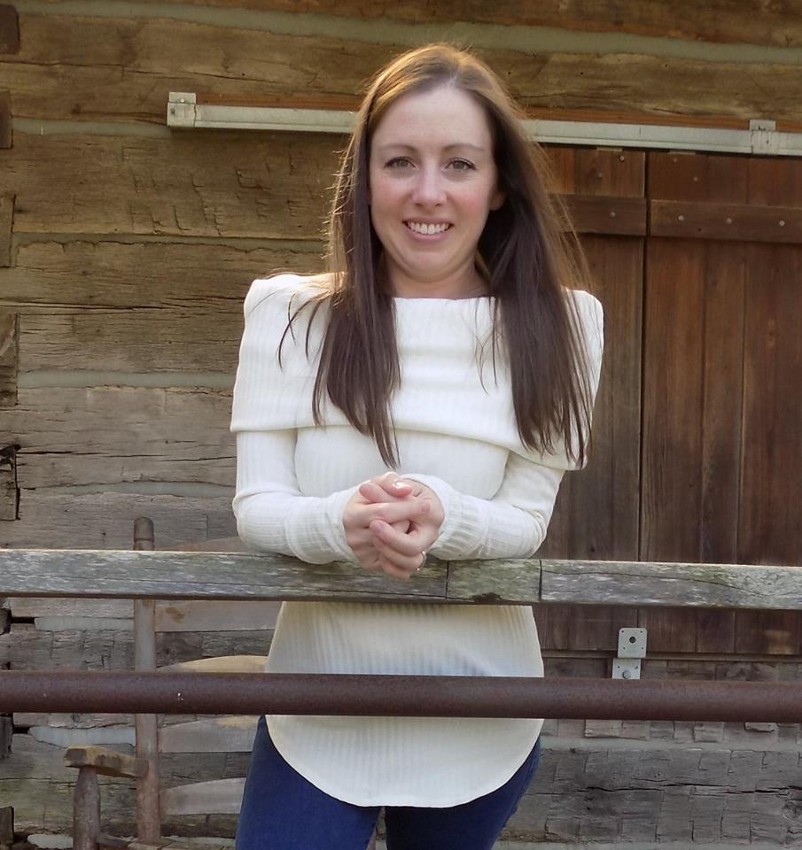 A woman with straight brown hair standing outside of a cabin, leaning on a railing, wearing a gorgeous white lightweight sweater with off-shoulder cowl and long cuffed sleeves. Hem of the sweater is hemmed and curved. Made using Ellie and Mac Sewing Patterns.