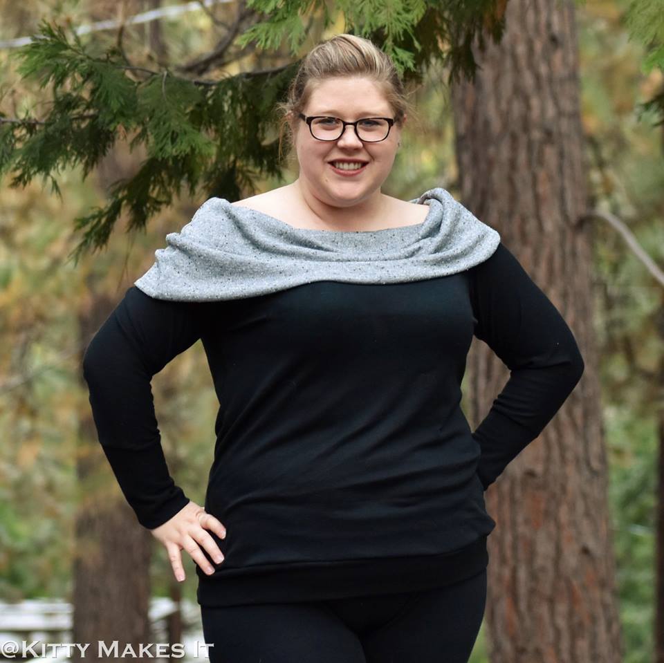 A woman with a ponytail and glasses wearing a black sweater with a grey slouchy cowl neck. 